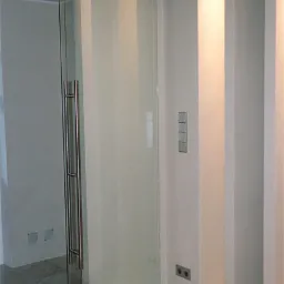 Szklane drzwi przesuwne z satynowym wykończeniem, zawieszone na metalowej szynie z widocznymi rolkami, z długim, pionowym uchwytem ze stali nierdzewnej, na tle białej ściany z włącznikami...