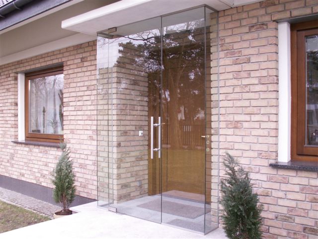 Szklana zabudowa wejścia do domu z cegły, widoczne drzwi z hartowanego szkła z długimi, pionowymi uchwytami.