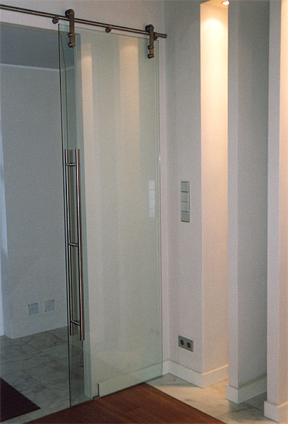 Szklane drzwi przesuwne z satynowym wykończeniem, zawieszone na metalowej szynie z widocznymi rolkami, z długim, pionowym uchwytem ze stali nierdzewnej, na tle białej ściany z włącznikami...