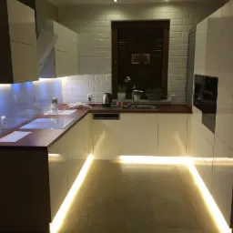 Nowoczesna kuchnia na wymiar z białymi frontami, drewnianym blatem i subtelnym oświetleniem LED pod szafkami, podkreślającym wzór płytek na podłodze.