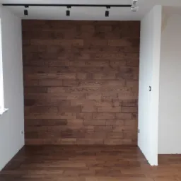 Wnętrze pokoju z drewnianą podłogą i ścianą z paneli imitujących drewno, widok na okno z widokiem na miasto, surowe wykończenie ścian.