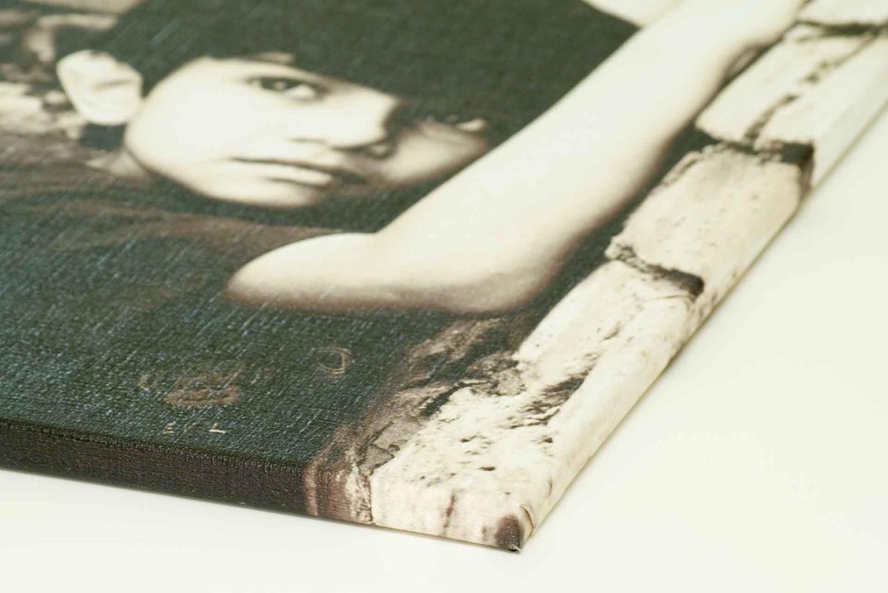 Detal czarno-białego portretu chłopca wydrukowanego na płótnie, widoczny fragment twarzy, ramienia i tła imitującego mur z kamieni, z ostrym kątem widzenia na krawędź płótna.