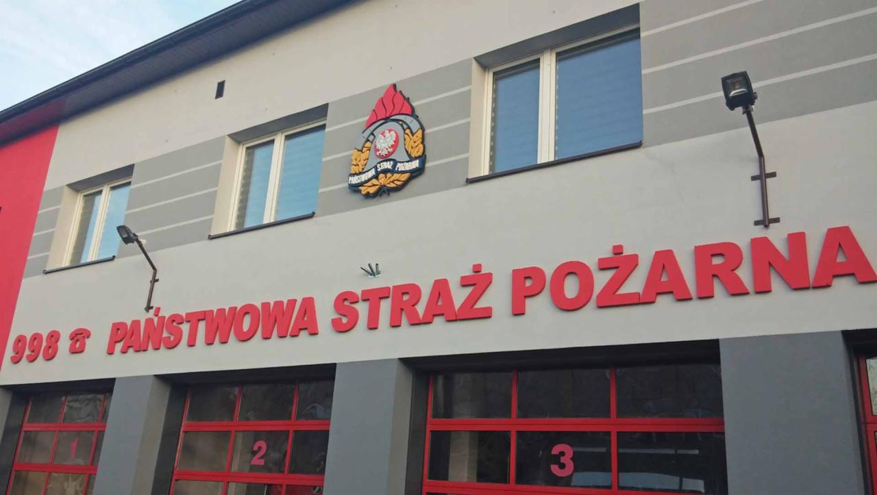 Fasada budynku Państwowej Straży Pożarnej z czerwonymi literami i symbolem strażackim nad garażami.