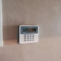 Biały panel sterowania alarmu firmy Salef Perfecto zamontowany na szarej, surowej ścianie, obok biała skrzynka elektryczna.