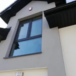 Zewnętrzny widok domu z zamontowanym czujnikiem alarmowym (czerwony kwadrat) pod dachem i oknem z odbijającym się niebem.