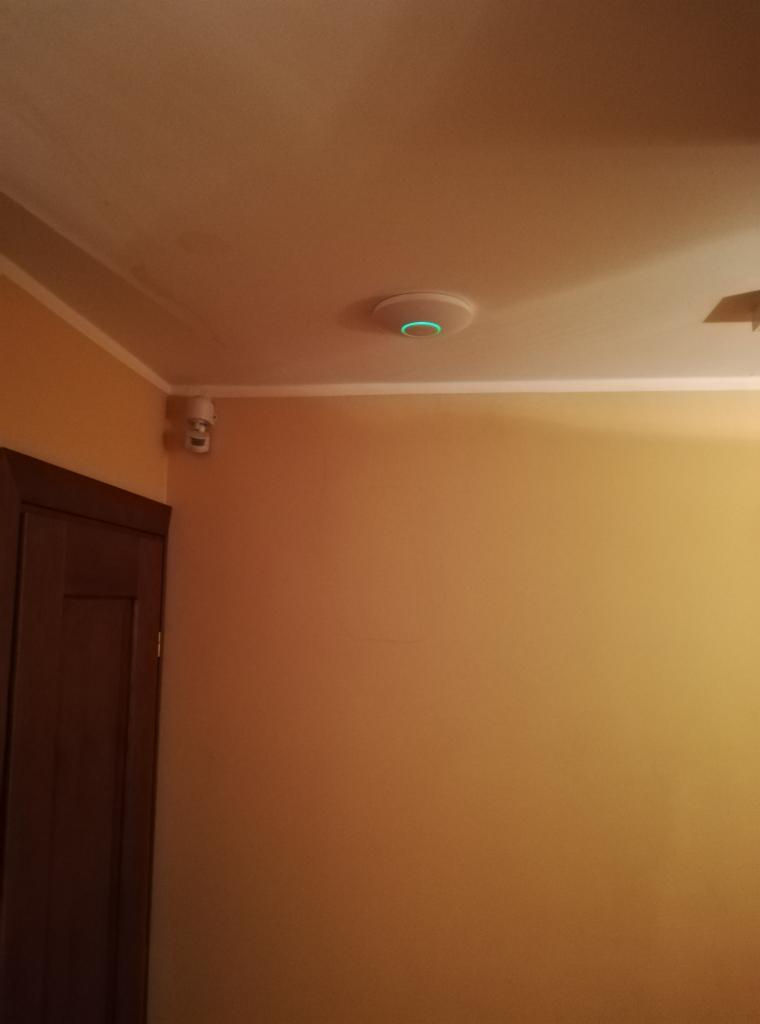 Biały, okrągły czujnik z zieloną diodą na suficie w pokoju z pomarańczowymi ścianami; kamera monitoringu zamontowana w rogu ściany obok brązowych drzwi.