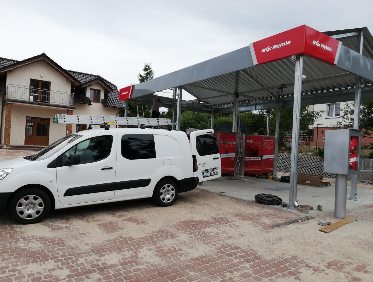 Biały samochód dostawczy z drabiną na dachu zaparkowany przed nowo wybudowaną samoobsługową myjnią samochodową z czerwonymi panelami.