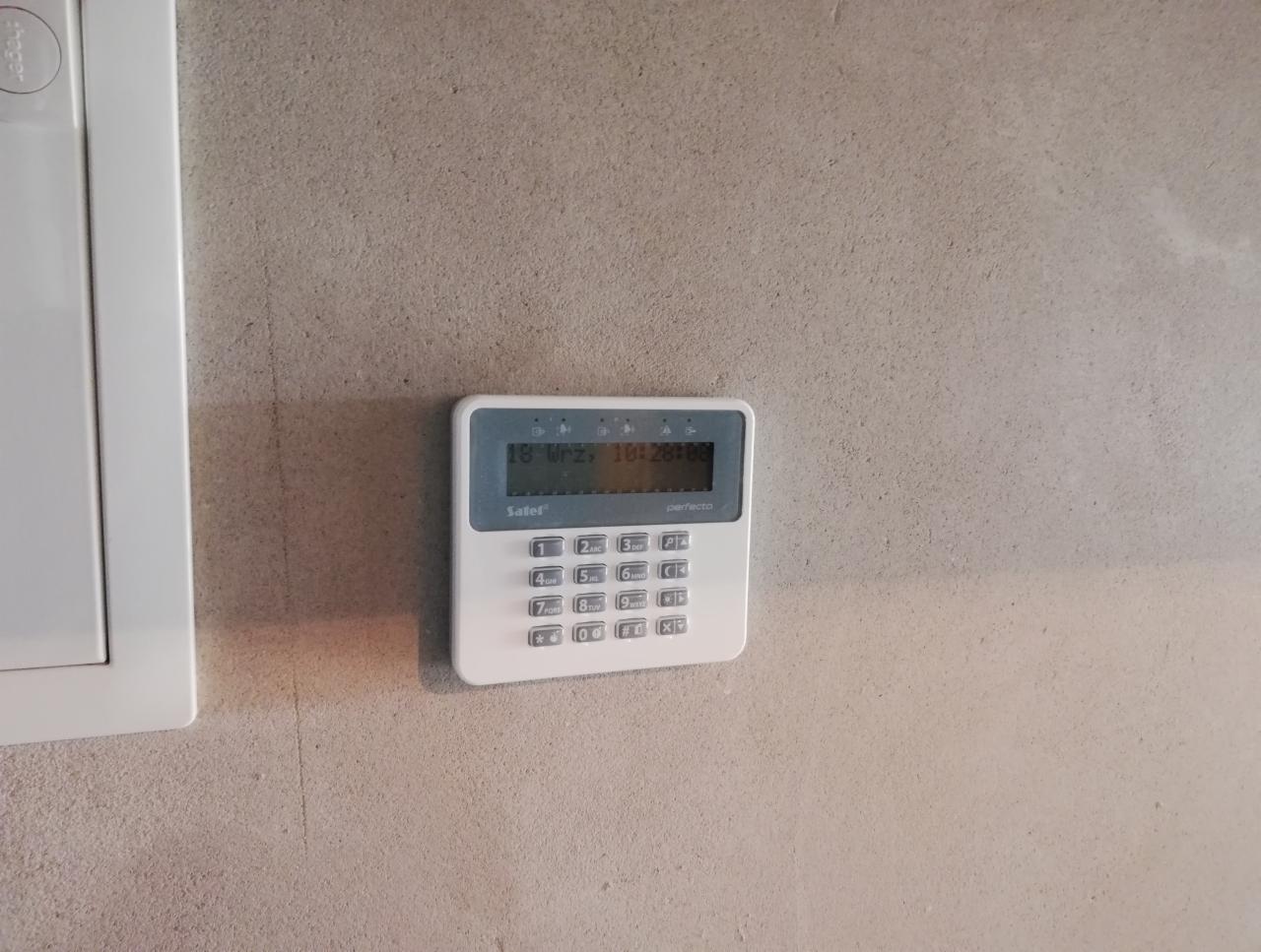 Biały panel sterowania alarmu firmy Salef Perfecto zamontowany na szarej, surowej ścianie, obok biała skrzynka elektryczna.