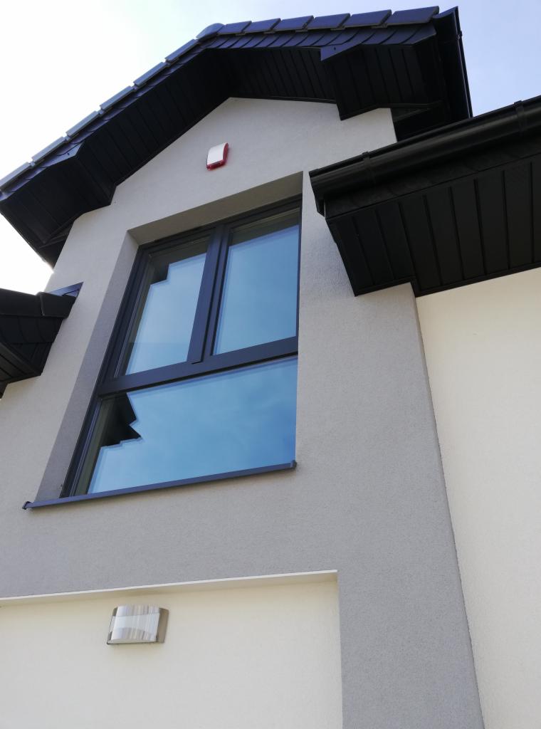 Zewnętrzny widok domu z zamontowanym czujnikiem alarmowym (czerwony kwadrat) pod dachem i oknem z odbijającym się niebem.