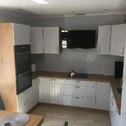 Jasna kuchnia z białymi szafkami i drewnianym blatem, widoczny czajnik na płycie grzewczej i stół z krzesłami w tle.