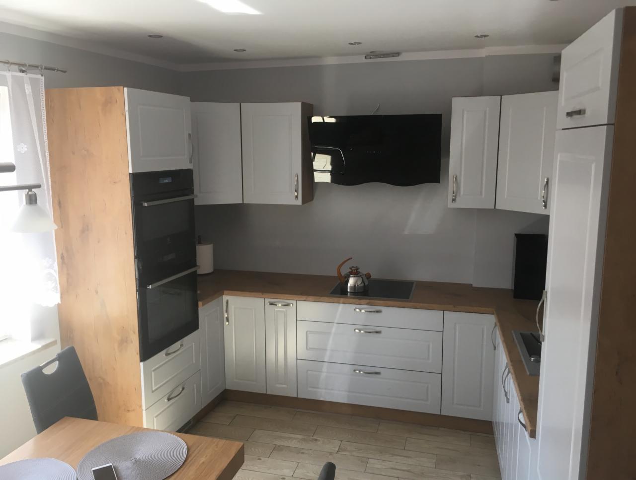 Jasna kuchnia z białymi szafkami i drewnianym blatem, widoczny czajnik na płycie grzewczej i stół z krzesłami w tle.