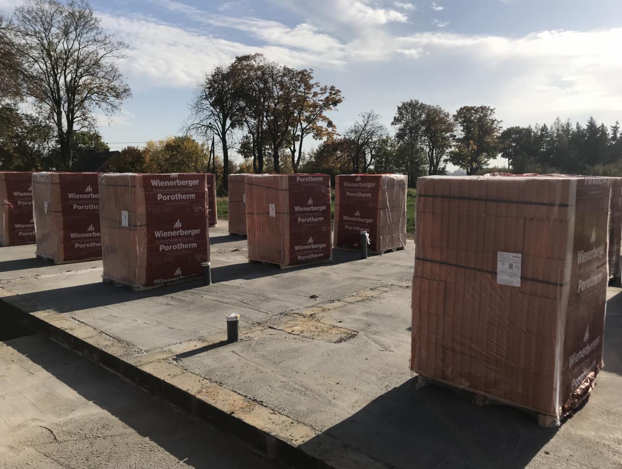 Palety z cegłami Porotherm Wienerberger na betonowym fundamencie budynku w trakcie budowy, widoczne drzewa w tle.