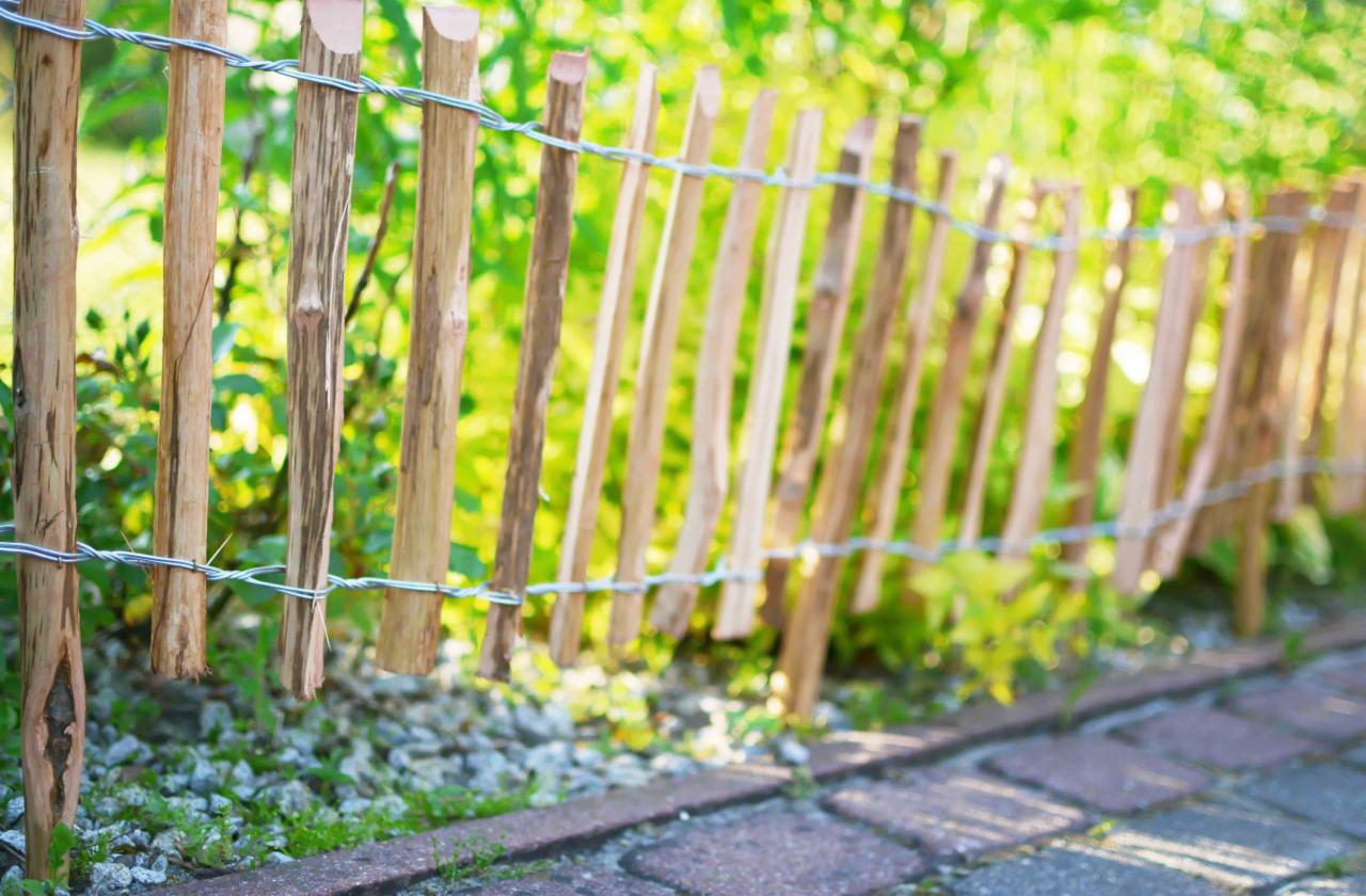 Niski płotek z surowego drewna, połączony drutem, oddzielający trawnik od ścieżki z kostki brukowej, z zielenią w tle.