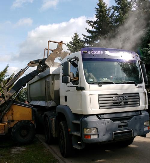 Żółta ładowarka z łyżką załadowuje gruz do białej ciężarówki MAN na tle drzew i błękitnego nieba.
