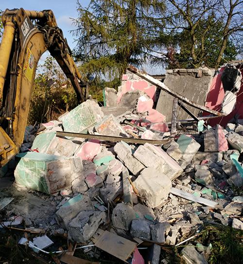 Żółta koparka CAT częściowo widoczna obok sterty gruzu z resztkami różowej i zielonej farby po zburzonym budynku na tle drzew