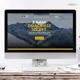 Ekran komputera iMac wyświetlający stronę internetową z motywem górskim i hasłem 'Z nami osiągniesz szczyt', na biurku z białą klawiaturą i myszką.
