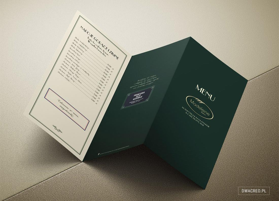 Otwarta karta menu restauracji w kolorze butelkowej zieleni z logo 'Madeleine Bakery' i listą napojów, leżąca na teksturowanym tle.