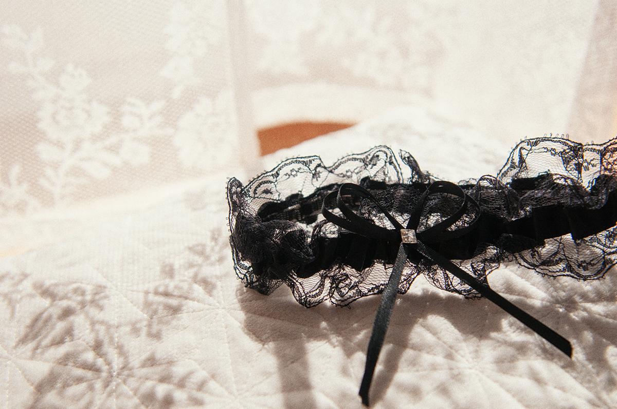 Czarna koronkowa podwiązka z kokardką i drobnym kamieniem na białej, pikowanej tkaninie z wzorem kwiatowym w tle.