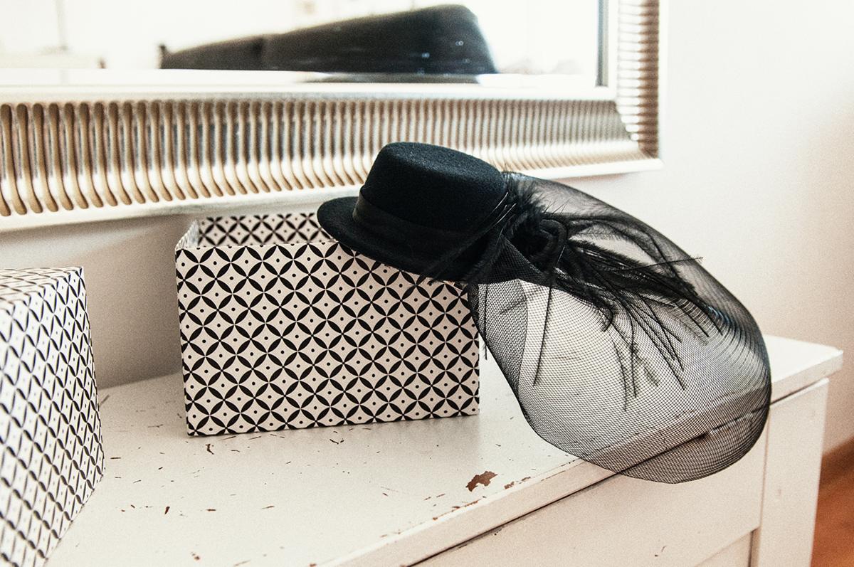 Czarny cylinder z woalką i piórami leży na pudełku z geometrycznym wzorem, obok drugiego pudełka, na białej komodzie pod lustrem w ozdobnej ramie.
