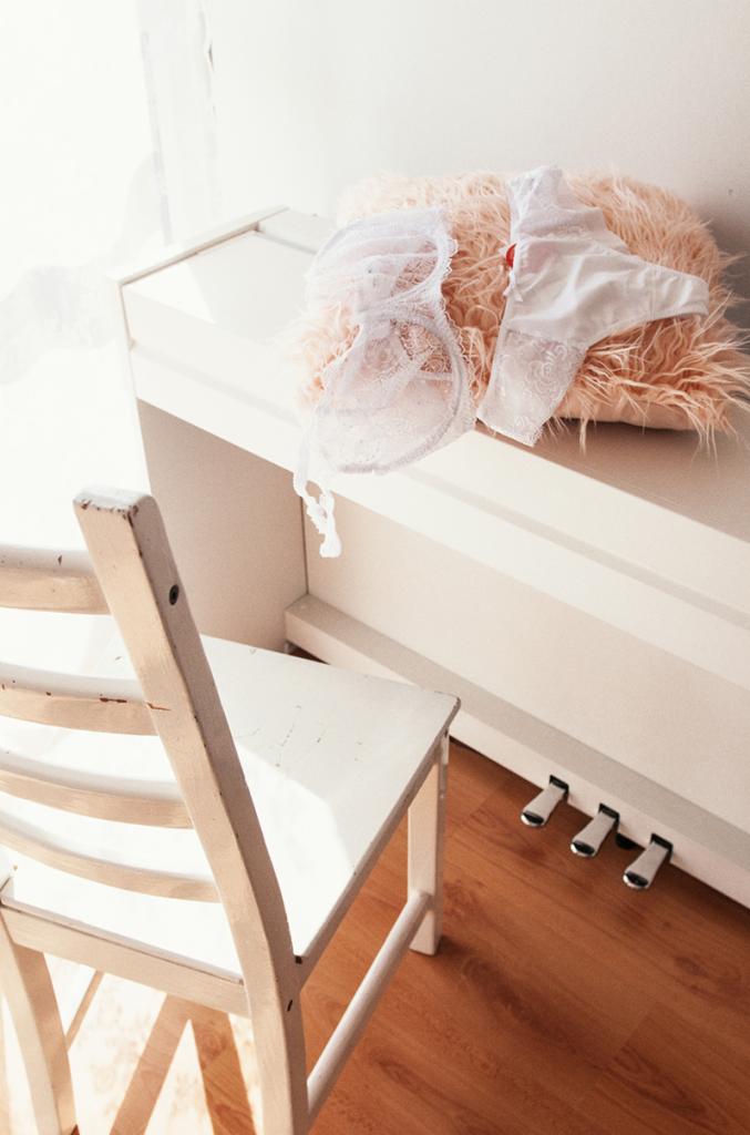 Biała, koronkowa bielizna leży na puszystej poduszce na białym pianinie, obok stoi białe krzesło z widocznymi śladami użytkowania.