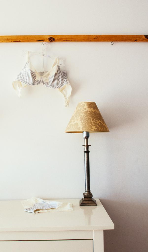 Stylizacja wnętrza z lampką z beżowym abażurem w kwiatowy wzór, biustonoszem w biało-szare paski wiszącym na wieszaku i dopasowaną chusteczką na blacie komody.