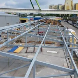 Montaż stalowej konstrukcji dachu hali przemysłowej z użyciem dźwigu, widok z góry na częściowo zadaszoną konstrukcję i okoliczne budynki.