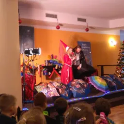 Animatorzy dla dzieci podczas występu na scenie z rekwizytami, w tle choinka bożonarodzeniowa i dziecięca publiczność.