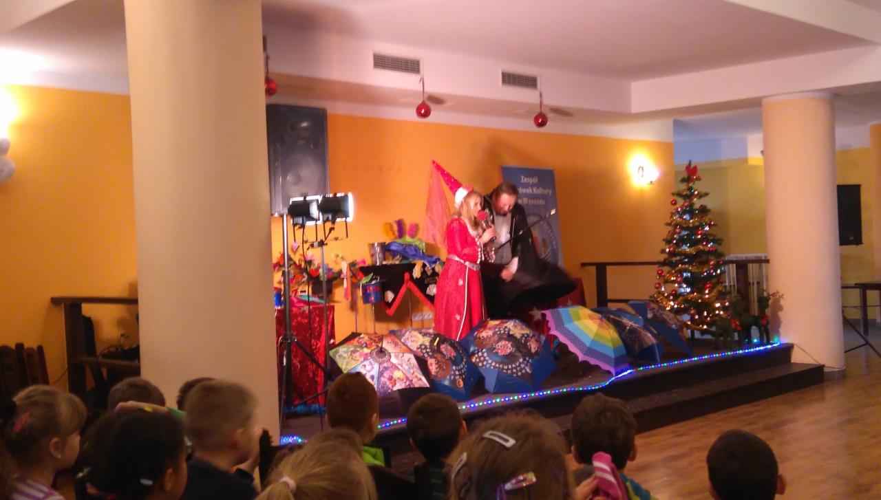 Animatorzy dla dzieci podczas występu na scenie z rekwizytami, w tle choinka bożonarodzeniowa i dziecięca publiczność.