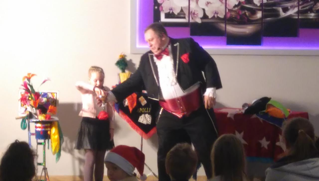 Iluzjonista w czarnym fraku z czerwoną muchą i pasem prezentuje sztuczkę z czerwoną kulką dziewczynce na scenie podczas występu dla dzieci.