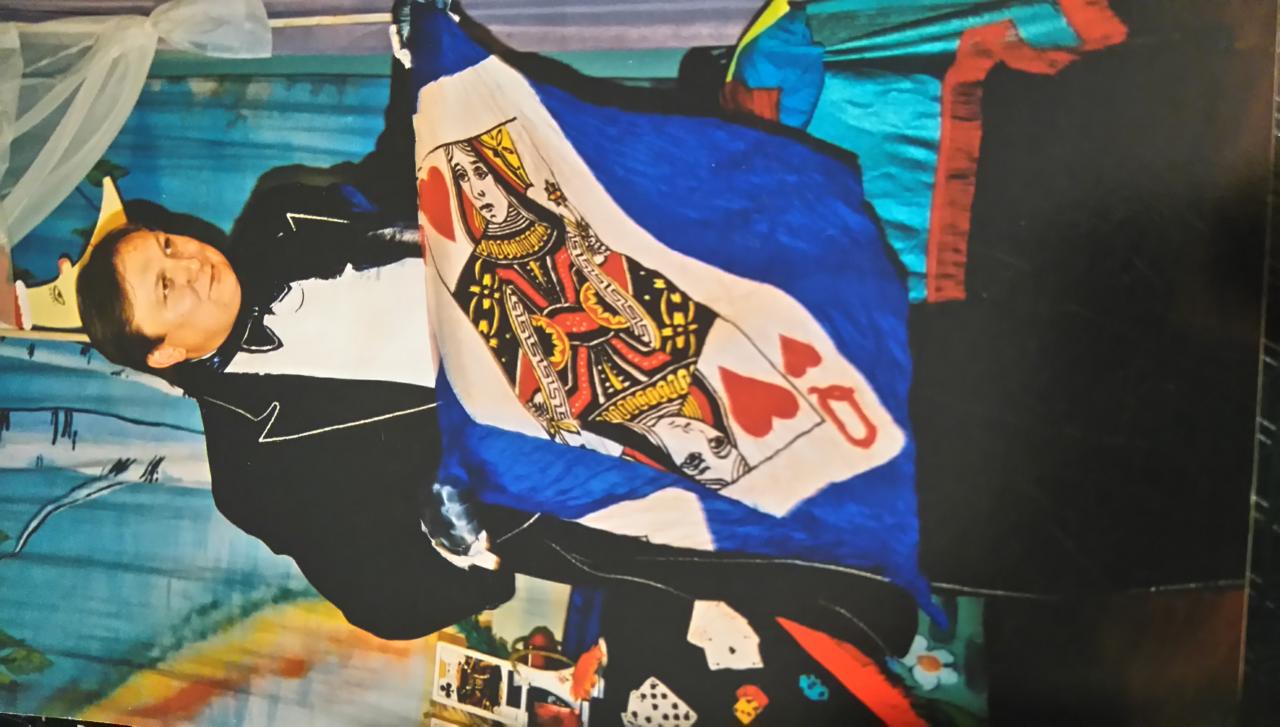 Mężczyzna w smokingu trzymający dużą kartę do gry z wizerunkiem Królowej Kier na tle sceny z rekwizytami.