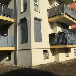 Nowoczesny budynek wielorodzinny z szarymi roletami zewnętrznymi opuszczonymi w oknach, balkony z szarą balustradą i żółtymi elementami, widok z dołu.