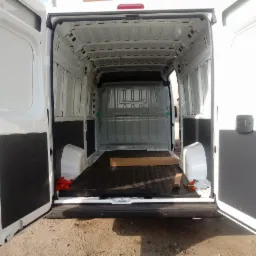 Wnętrze białego busa z otwartymi drzwiami, pusta przestrzeń ładunkowa wyłożona gumową matą, widoczne pasy transportowe i drewniane elementy zabezpieczające.