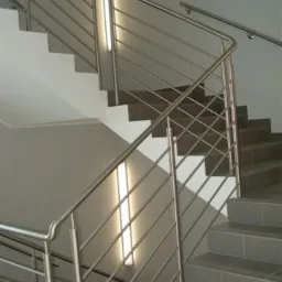 Schody wyłożone szarą płytką z nowoczesną balustradą ze stali nierdzewnej, oświetlone pionową lampą ścienną.