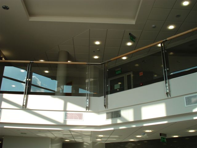 Szklana balustrada z drewnianym poręczą w nowoczesnym budynku, widok z dołu na górny poziom z widocznymi oknami i sufitem z oświetleniem punktowym.