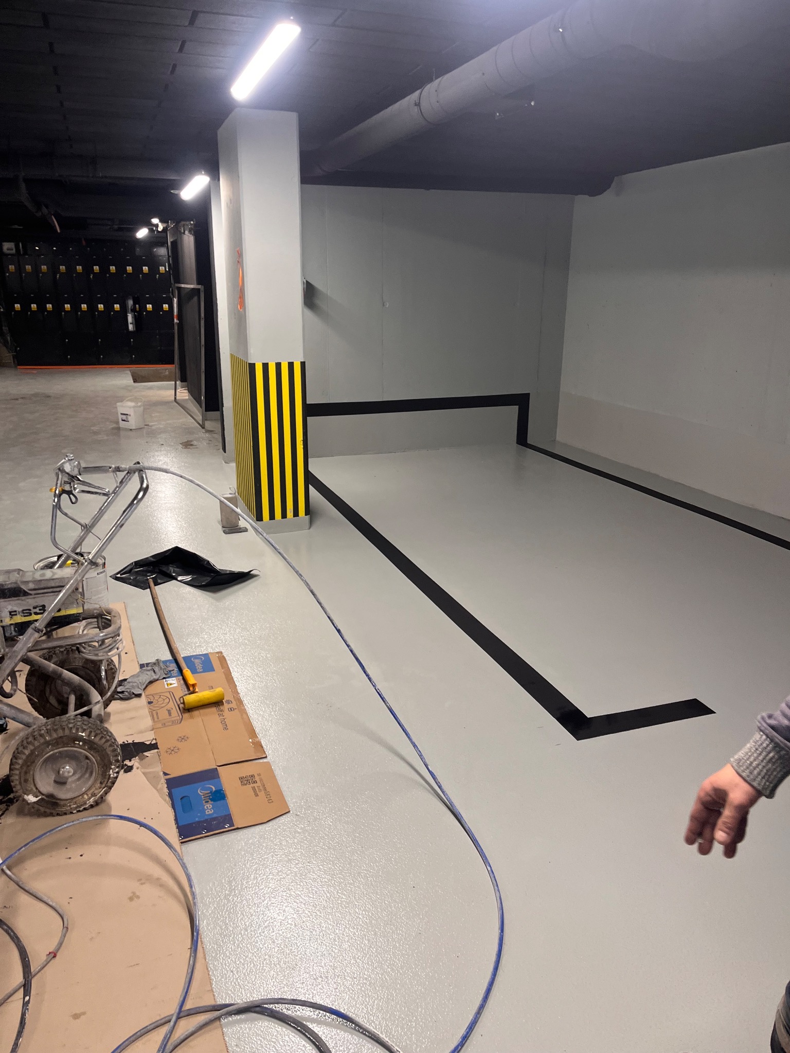 Szara posadzka żywiczna z czarnymi liniami w garażu podziemnym. Widoczny sprzęt do malowania, kolumna w czarno-żółte pasy i fragment ręki. Oświetlenie sufitowe.