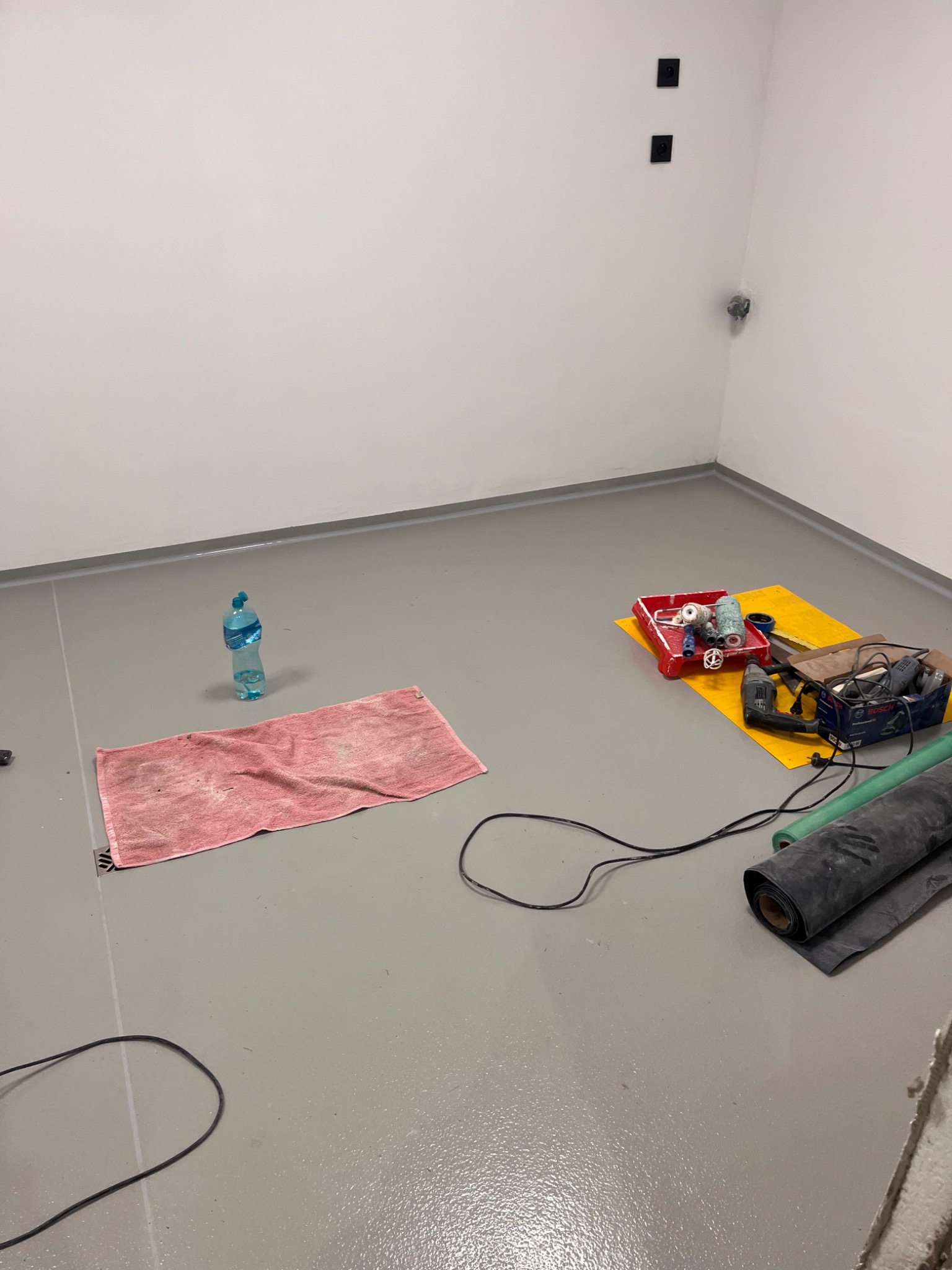 Jasnoszara posadzka żywiczna w trakcie realizacji, z narzędziami, butelką wody i ręcznikiem. Widoczne czarne gniazdka elektryczne na białej ścianie.