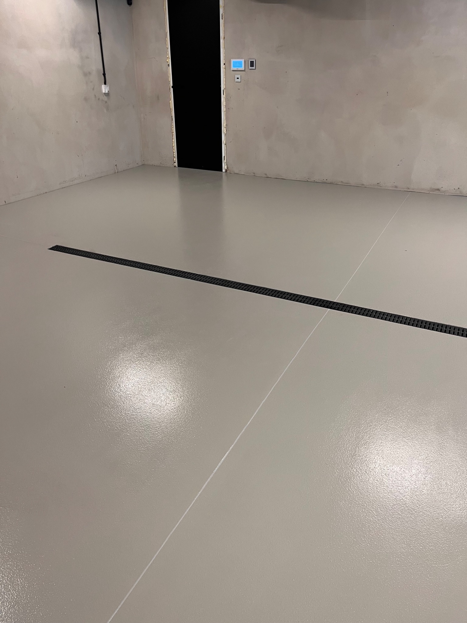 Jasnoszara, gładka posadzka żywiczna z liniowym odpływem w nowym pomieszczeniu. Ściany surowe, betonowe. Widoczne elementy instalacji elektrycznej.