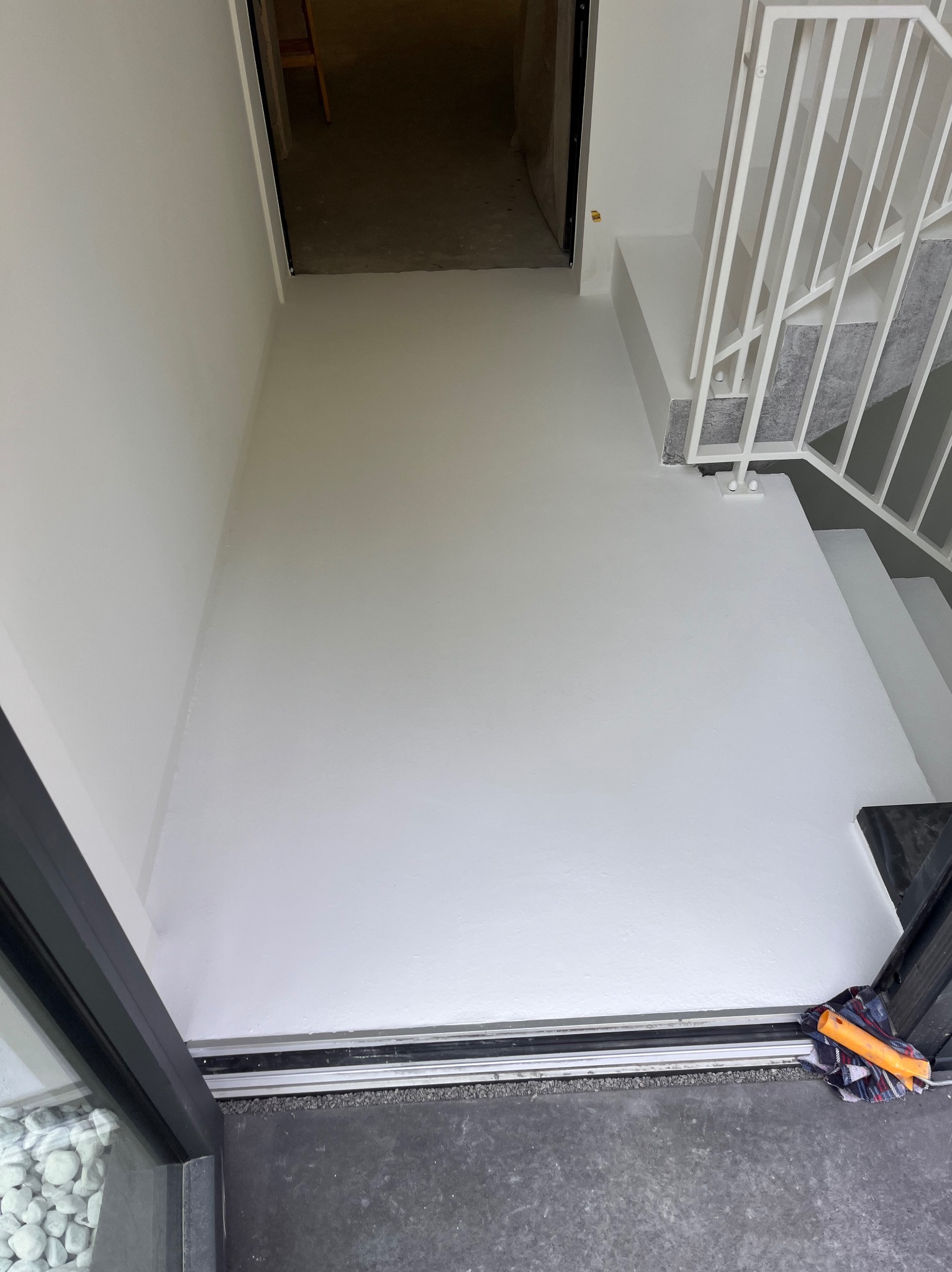 Biała posadzka żywiczna w korytarzu z fragmentem schodów i wejściem. Widoczny fragment okna z kamieniami dekoracyjnymi i narzędzie pracy na podłodze.