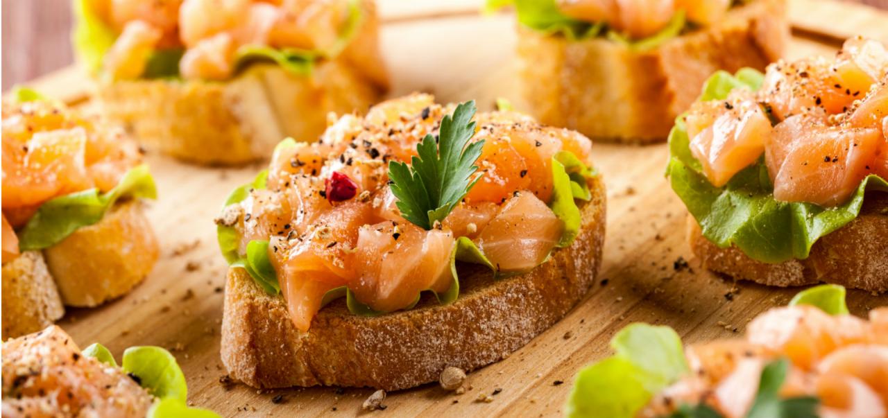 Apetyczne kanapeczki z łososiem, świeżą sałatą i przyprawami, ułożone na drewnianej desce, z bliska widoczny listek pietruszki i czerwony pieprz.