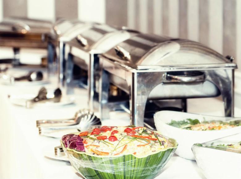 Bufet z podgrzewaczami, sałatkami w dekoracyjnych misach i szczypcami do nakładania, ustawiony na białym stole z szarym, pasiakowym tłem.