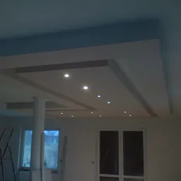 Wykończony sufit podwieszany z oświetleniem punktowym i kolumną nośną, widoczna drabina malarska w rogu pokoju.