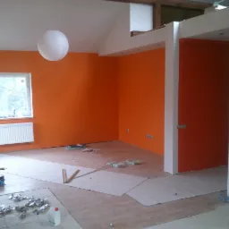 Wnętrze w trakcie malowania na pomarańczowo, z widocznymi zabezpieczeniami podłogi, wiszącą lampą kulą i fragmentem okna z grzejnikiem.