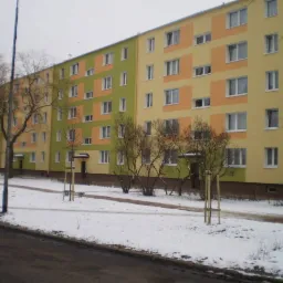 Wielopiętrowy budynek mieszkalny po dociepleniu elewacji, z zastosowaniem kolorowych paneli w odcieniach żółci, pomarańczy i zieleni, widziany w zimowej scenerii z resztkami śniegu na chodniku.