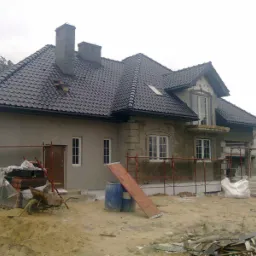 Dom jednorodzinny w trakcie prac elewacyjnych: widoczne rusztowania, częściowo otynkowana ściana, przygotowane materiały budowlane i stos płytek ceramicznych Koramic.