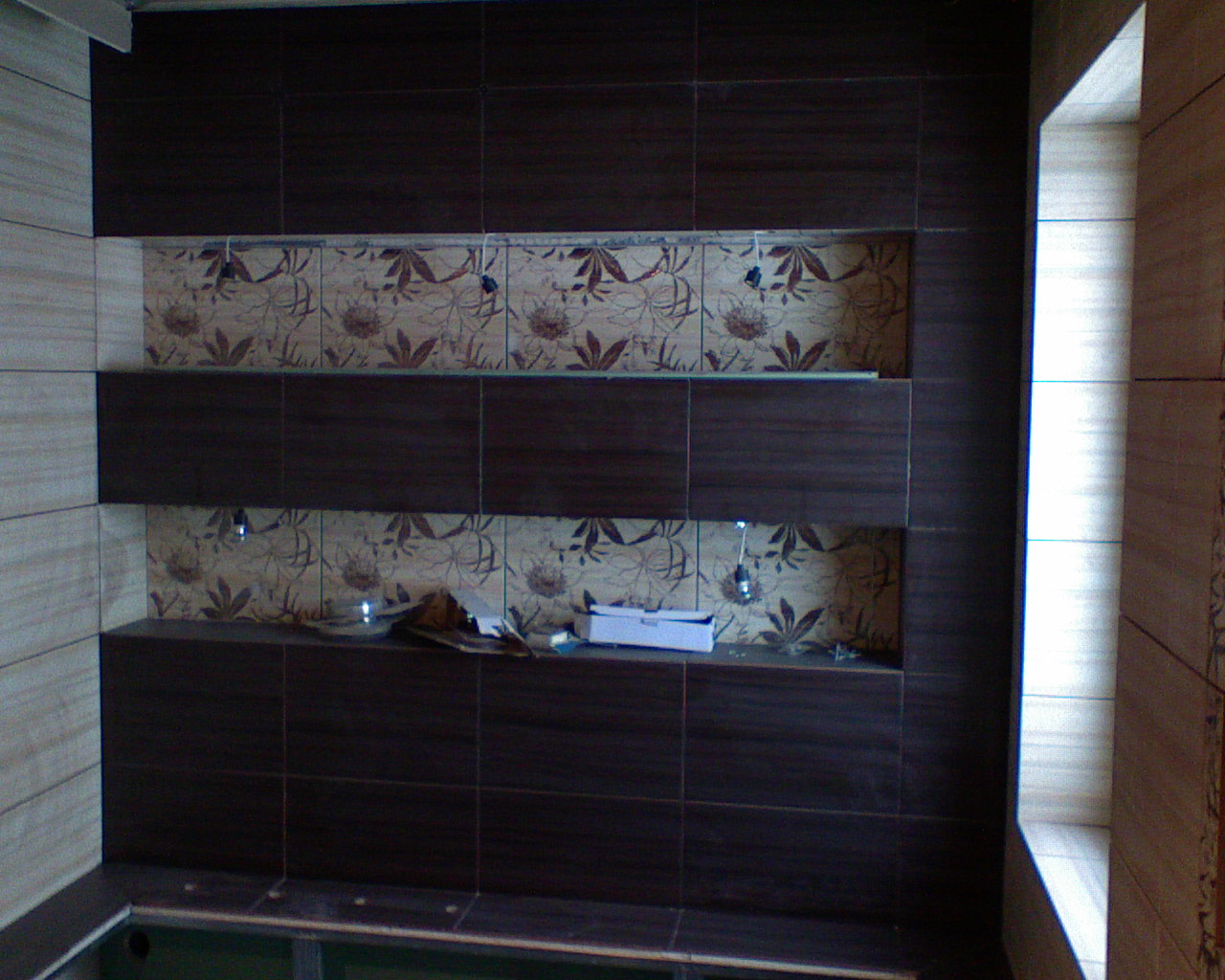 Ściana łazienki wyłożona ciemnobrązowymi i beżowymi płytkami z motywem roślinnym, z wnękami oświetlonymi małymi lampkami, w trakcie układania płytek widoczne narzędzia i pudełko.