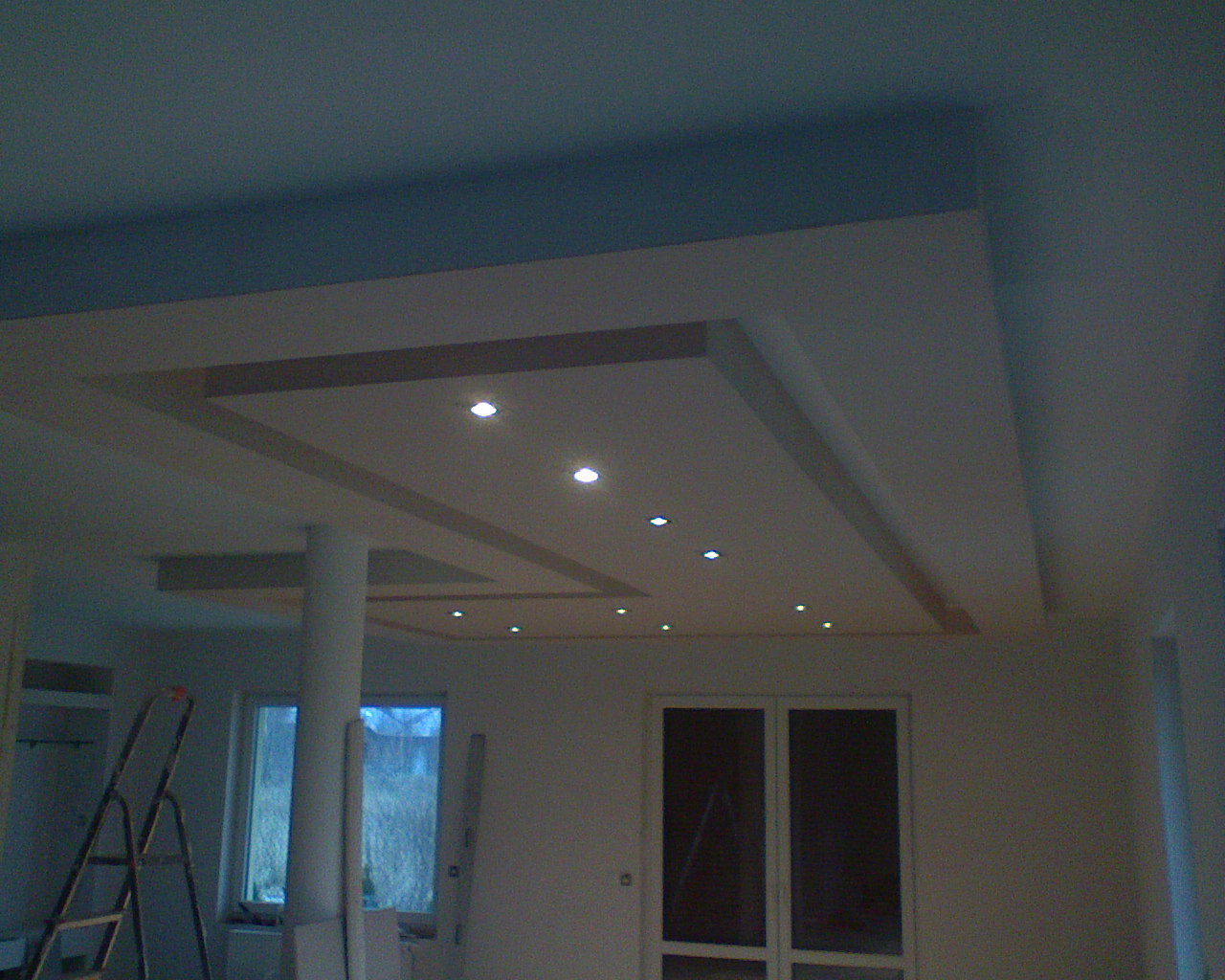 Wykończony sufit podwieszany z oświetleniem punktowym i kolumną nośną, widoczna drabina malarska w rogu pokoju.