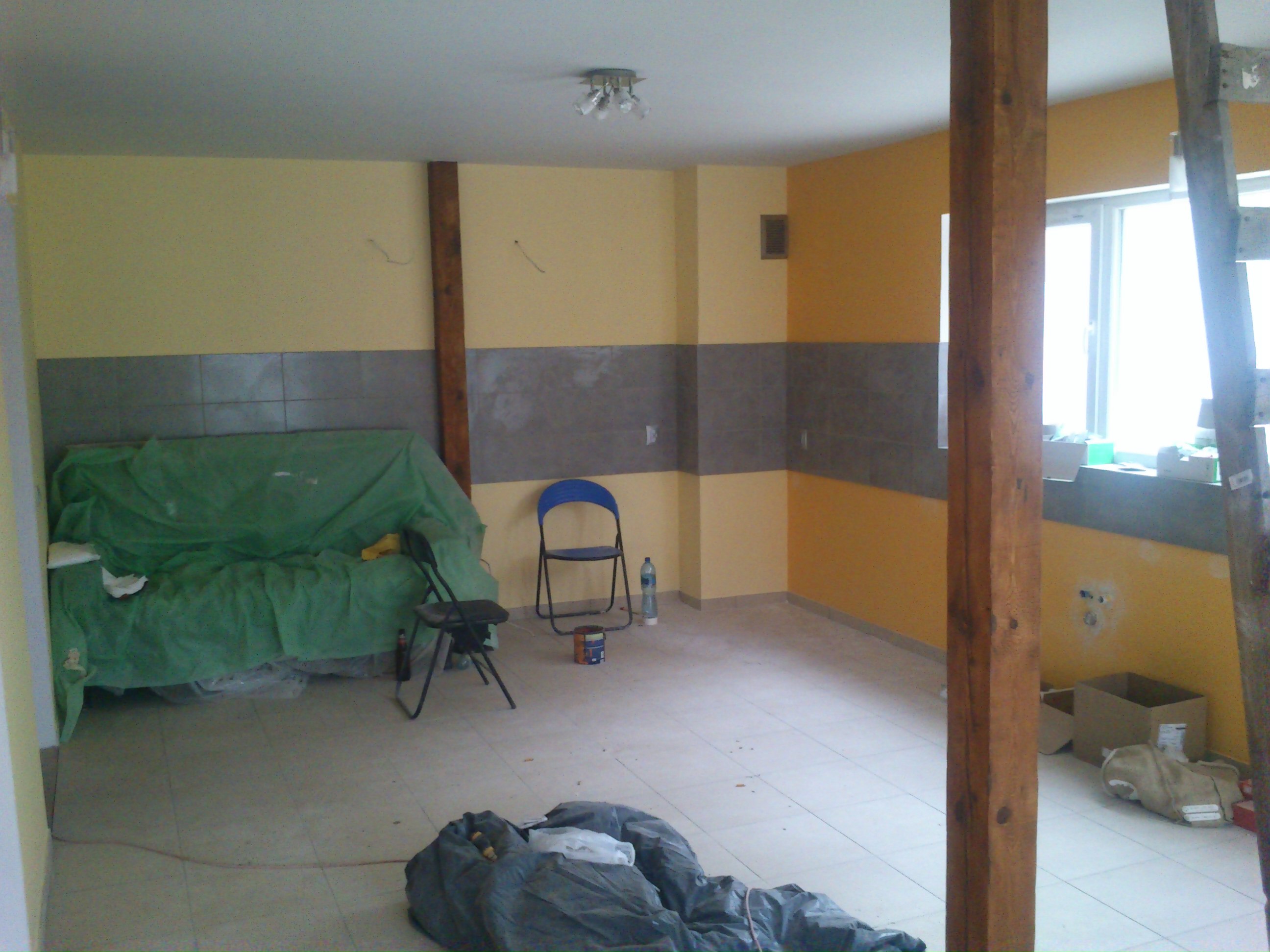 Pokój w trakcie remontu: ściany pomalowane częściowo na żółto i szaro, widoczne drewniane filary, drabina oparta o okno, podłoga wyłożona jasnymi płytkami, meble przykryte folią ochronną, butelka...