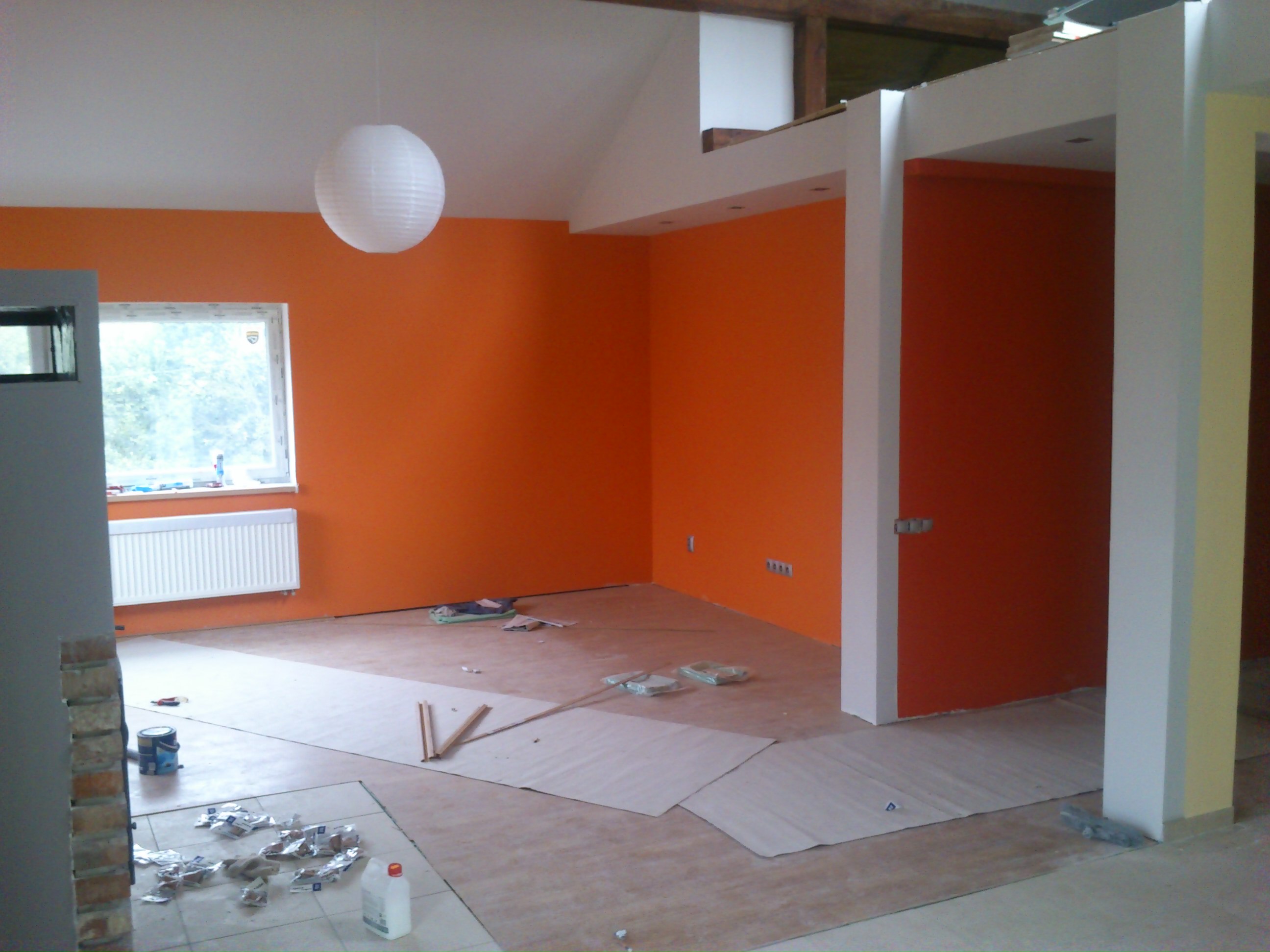 Wnętrze w trakcie malowania na pomarańczowo, z widocznymi zabezpieczeniami podłogi, wiszącą lampą kulą i fragmentem okna z grzejnikiem.