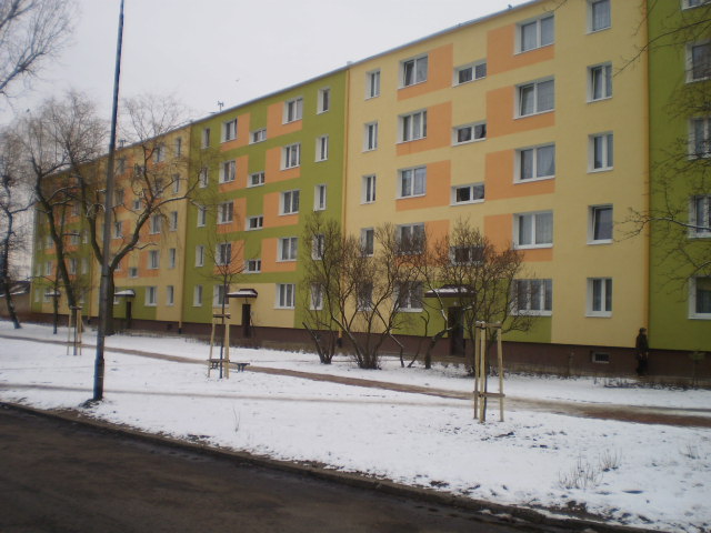 Wielopiętrowy budynek mieszkalny po dociepleniu elewacji, z zastosowaniem kolorowych paneli w odcieniach żółci, pomarańczy i zieleni, widziany w zimowej scenerii z resztkami śniegu na chodniku.