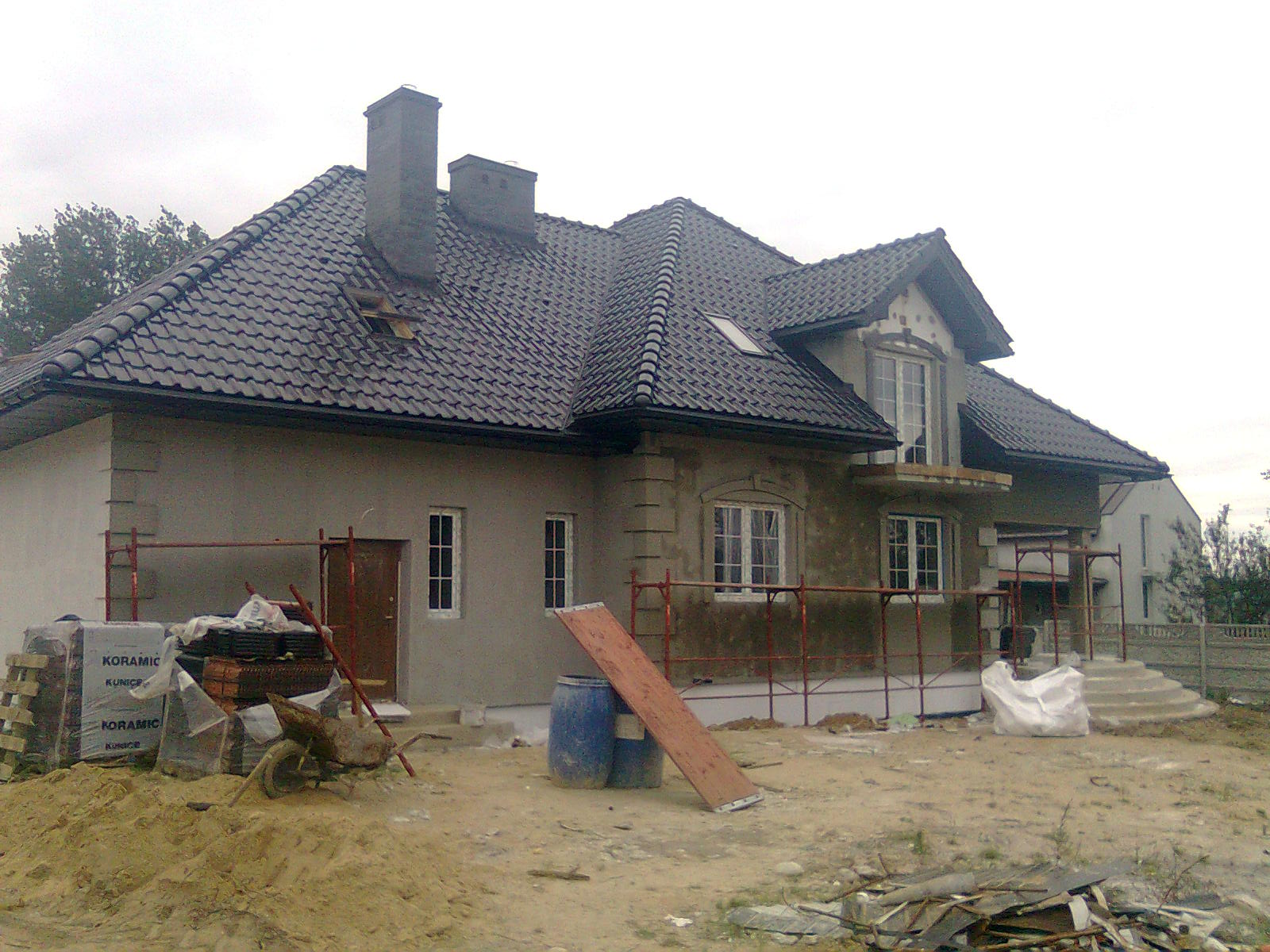 Dom jednorodzinny w trakcie prac elewacyjnych: widoczne rusztowania, częściowo otynkowana ściana, przygotowane materiały budowlane i stos płytek ceramicznych Koramic.